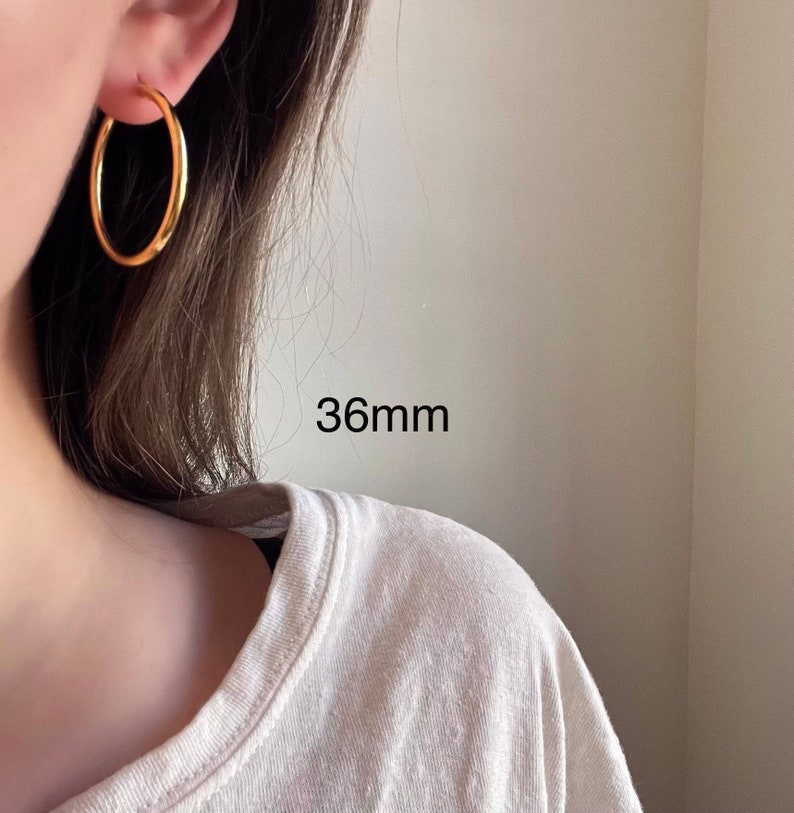 以下が含まれることがあります： 直径36mmのゴールドフープピアス。ピアスは人の耳につけられています。