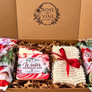 May include: A cardboard gift box containing a bar of Rose & Vine Naturals Winter Wonderland Cranberry Aspen soap with red and white swirls, and a beige crocheted washcloth tied with a red ribbon, nestled in brown paper crinkle fill.