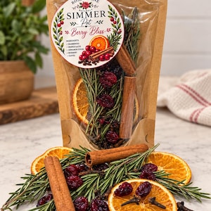 May include: A clear plastic pouch with a kraft paper backing containing dried orange slices, cinnamon sticks, rosemary sprigs, cranberries, and cloves. The label reads Rose & Vine Naturals Simmer Pot Berry Bliss with a list of ingredients.