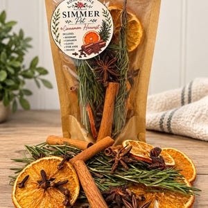 May include: A brown paper pouch labeled Rose & Vine Naturals Simmer Pot Cinnamon Harvest containing dried rosemary, cinnamon sticks, star anise, cloves, and orange slices. Loose ingredients are scattered in front of the pouch on a wooden surface.