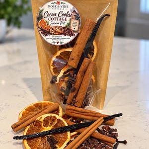 May include: A brown paper pouch containing a Cocoa Cookie Cottage Simmer Pot mix. The label features text and illustrations of cookies and oranges. Dried orange slices, cinnamon sticks, vanilla beans, and cocoa nibs are visible inside.