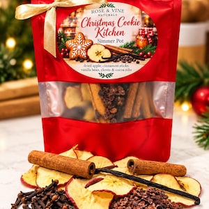 May include: A red pouch labeled Rose & Vine Naturals Christmas Cookie Kitchen Simmer Pot, containing dried apple, cinnamon sticks, vanilla bean, cloves, and cocoa nibs. A gold ribbon is tied at the top, and the ingredients are displayed in front.