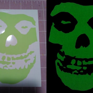 May include: A green glow-in-the-dark skull sticker with a white outline. The skull has a wide smile with teeth and closed eyes.