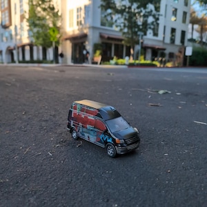 May include: A black toy van with a colorful graffiti design on the side and roof. The van has silver rims and is parked on a dark asphalt surface. The background shows a blurred view of buildings and trees.