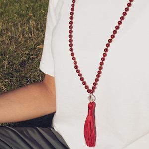 Peut inclure: Un long collier de perles rouges avec un pompon. Le collier est composé de petites perles rondes et d'une perle de cristal transparente. Le pompon est de couleur rouge foncé. Le collier est porté sur une chemise blanche.