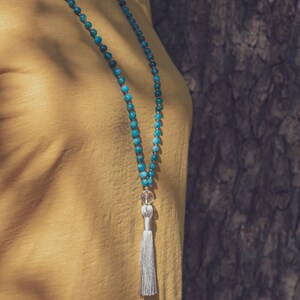 Peut inclure: Un long collier composé de perles turquoise, d'un cristal transparent et d'un pompon argenté. Le collier est présenté sur un vêtement jaune et un fond brun texturé.