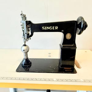 Puede incluir: Máquina de coser Singer negra con letras doradas, colocada sobre una mesa blanca. La máquina presenta un gran volante, un conjunto de agujas plateadas y una base redonda. El logotipo de Singer es visible.
