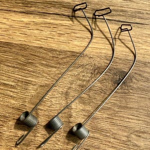 以下が含まれることがあります： ループ状の端とコイル状のベースを備えた、3つの銀色の金属スプリング。スプリングは湾曲したデザインです。背景は木製の表面です。