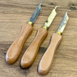 Könnte beinhalten: Drei Schnitzmesser mit Holzgriffen und gebogenen Klingen. Die Klingen sind silberfarben und die Griffe hellbraun. Die Messer haben messingfarbene Kragen, wo die Klinge auf den Griff trifft. Die Messer sind diagonal auf einer Holzoberfläche angeordnet.