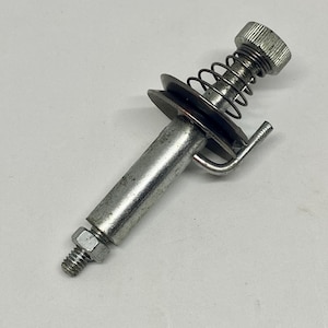 May include: A metal spring-loaded pin assembly with a knurled head, a spring, and a small pulley. The pin has a threaded end with a nut. The assembly is silver-colored and appears to be a mechanical component.