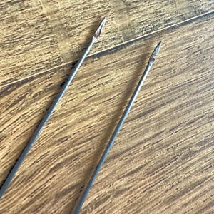 Könnte beinhalten: Zwei antike Metallpfeilspitzen mit spitzen Spitzen. Die Pfeilspitzen sind grau und haben ein schlankes, längliches Design. Der Hintergrund ist eine Holzoberfläche mit einem natürlichen Maserungsmuster.
