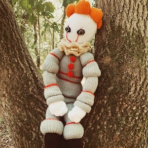 May include: A knitted doll of a clown with orange hair, white skin, and red and black clothing. The doll is sitting on a tree trunk.