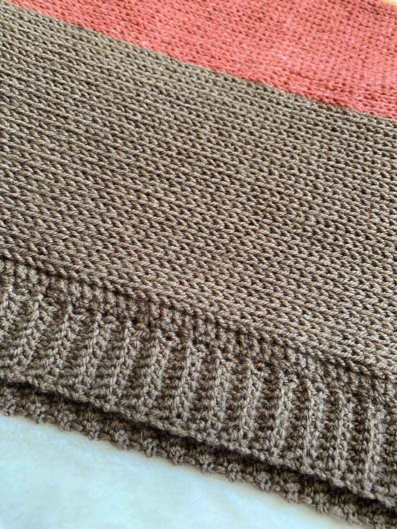 May include: A brown and coral striped knitted blanket with a textured weave. The blanket is folded in half and the edges are visible.