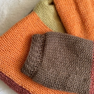May include: A striped knitted blanket in shades of brown, orange, and green. The blanket is folded in half, with the brown side facing up.