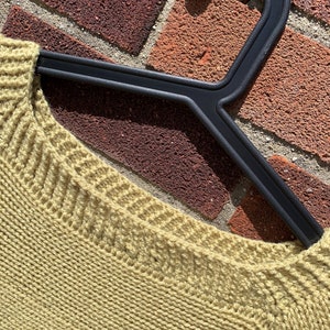 May include: A close-up of a light green knitted sweater with a ribbed neckline. The sweater is hanging on a black plastic hanger.