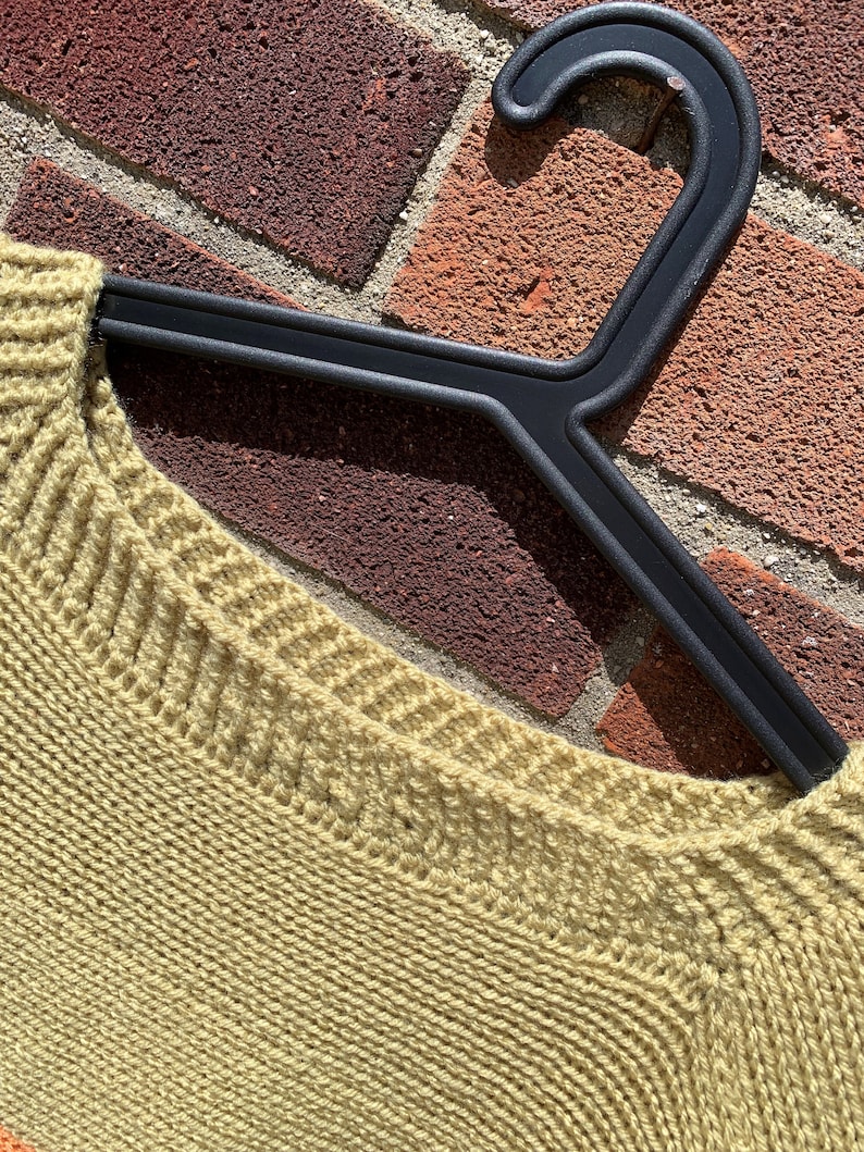 May include: A close-up of a light green knitted sweater with a ribbed neckline. The sweater is hanging on a black plastic hanger.
