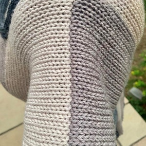 May include: Close-up of a knitted fabric with a gradient of colors, from light gray to dark gray. The fabric is made of soft yarn and has a textured surface. The fabric is folded over itself, creating a layered effect.