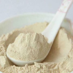 Puede incluir: Un bol de cerámica blanca lleno de un polvo beige claro. Una cuchara de cerámica blanca está parcialmente sumergida en el polvo, con el mango sobresaliendo del bol.