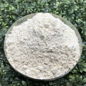 Può includere: Primo piano di una polvere bianca in un piatto di vetro trasparente. La polvere è fine e ha una consistenza leggermente granulosa. Il piatto è appoggiato su un letto di foglie verdi.