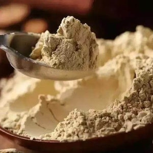 Puede incluir: Primer plano de una cuchara que recoge un montón de polvo marrón claro. El polvo está en un bol y tiene una textura rugosa.