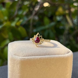May include: A gold ring with a red gemstone set in a delicate, ornate design. The ring is on a white velvet box.