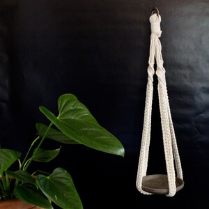Puede incluir: Un macetero de macramé blanco con una maceta redonda de cerámica marrón. El macetero está hecho de cuerda gruesa anudada y tiene un diseño simple.