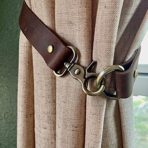 May include: A brown leather curtain tieback with a metal hook and ring. The tieback is wrapped around a beige linen curtain.