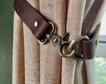May include: A dark brown leather curtain tieback featuring a brass-toned metal swivel lobster clasp and a circular ring. The leather strap is secured with small brass rivets, holding a textured, light beige fabric curtain in a gathered position.