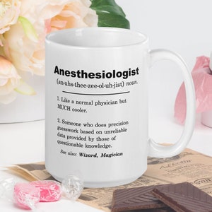 May include: White ceramic mug printed with the definition of "Anesthesiologist" in black. The definition includes the pronunciation, a humorous description, and related terms. The mug is on a table with flowers, sweets, and chocolate.
