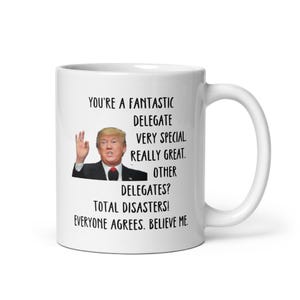 May include: White ceramic coffee mug with a black and white image of a man with a serious expression. The text on the mug reads: "You're a fantastic delegate. Very special. Really great. Other delegates? Total disasters! Everyone agrees. Believe me."