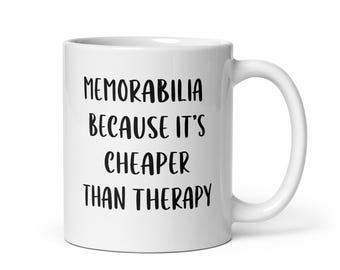 May include: White ceramic mug with the text "Memorabilia Because It's Cheaper Than Therapy" in black, bold font. The mug has a curved handle and a glossy finish, suitable for hot or cold beverages.