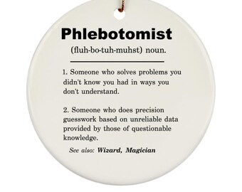 May include: White ceramic ornament with a black and white definition of the word "Phlebotomist" with the pronunciation "(fluh-bo-tuh-muhst)". The definition is: "1. Someone who solves problems you didn't know you had in ways you don't understand. 2. Someone who does precision guesswork based on unreliable data provided by those of questionable knowledge. See also: Wizard, Magician"