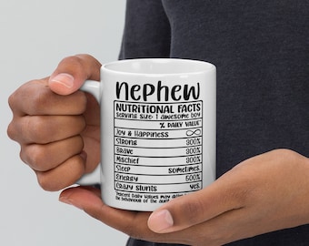May include: White ceramic mug with a black design that reads "nephew" and "Nutritional Facts" with a list of qualities and percentages. The mug is being held by a hand.
