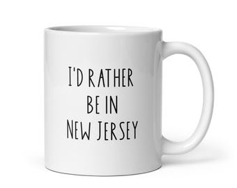 May include: White ceramic mug with the text "I'D RATHER BE IN NEW JERSEY" in black, sans-serif font. The mug has a curved handle and a glossy finish. A simple, everyday coffee mug.