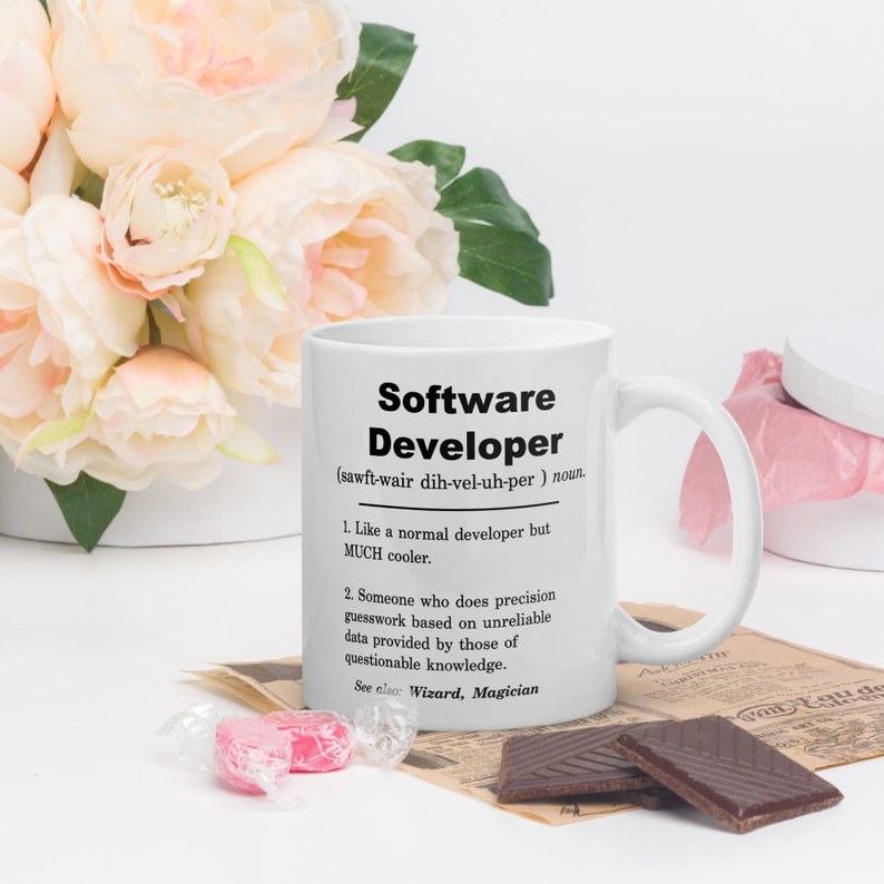 Op de afbeelding: Een witte keramische mok met een zwart ontwerp met de tekst "Software Developer (sawft-wair dih-vel-uh-per) noun. 1. Like a normal developer but MUCH cooler. 2. Someone who does precision guesswork based on unreliable data provided by those of questionable knowledge. See also: Wizard, Magician"