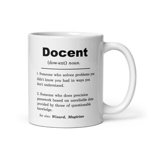 May include: White ceramic coffee mug with a black and white definition of the word "Docent" printed on it. The definition includes the pronunciation, "(dow-snt)", and two definitions. The mug has a black handle.