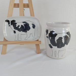 May include: A white ceramic dish and a jar, both adorned with a black and white cow design. The rectangular dish is on a wooden easel. The jar has a rounded shape with a wide opening.