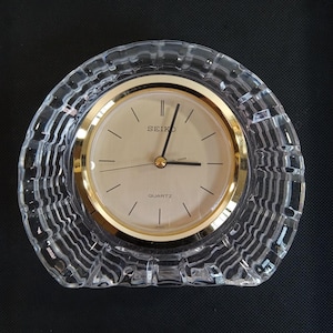 May include: A Seiko quartz clock with a gold-tone face and hands, set within a clear, textured crystal casing. The clock face has black markers and the word "SEIKO" at the top. The clock is a decorative item.