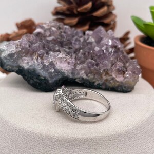 May include: A silver ring with a large, round, clear stone surrounded by smaller stones. The ring is set on a white surface with a purple amethyst crystal behind it.