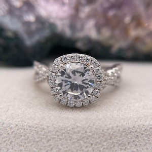 Puede incluir: Un anillo de compromiso de plata con una piedra central grande, redonda y transparente rodeada por un halo de piedras m&aacute;s peque&ntilde;as. La banda est&aacute; retorcida y presenta piedras m&aacute;s peque&ntilde;as adicionales.