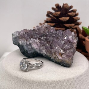 Puede incluir: Un anillo de compromiso de plata con una piedra central redonda y clara rodeada de piedras m&aacute;s peque&ntilde;as. El anillo tiene una banda retorcida y se muestra sobre una superficie blanca con un cristal de amatista p&uacute;rpura.