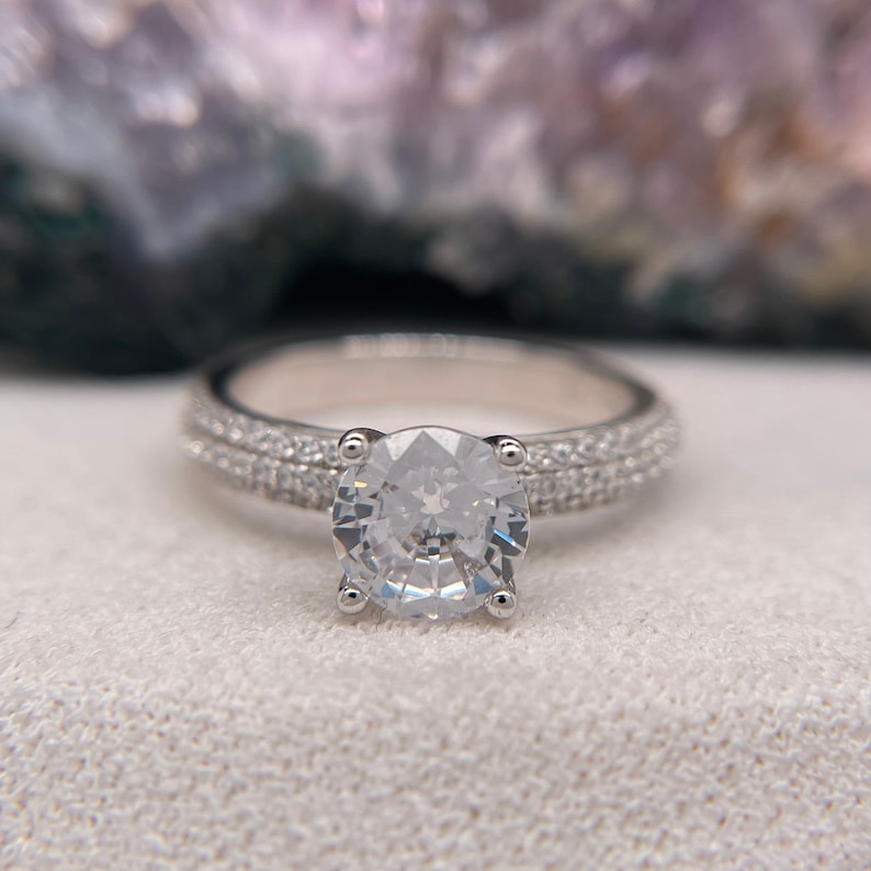 May include: A silver engagement ring with a large, round, clear centre stone and a double row of smaller stones around the band.