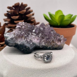 Puede incluir: Un anillo de compromiso de plata con una gran piedra redonda central rodeada de piedras m&aacute;s peque&ntilde;as. El anillo tiene una banda retorcida y est&aacute; colocado sobre una superficie blanca.