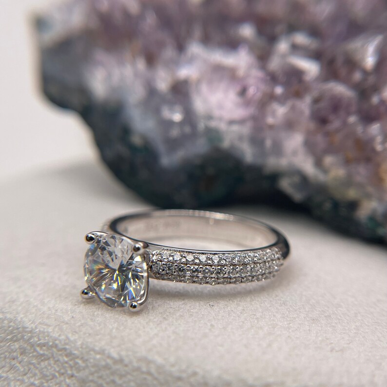May include: A silver engagement ring featuring a large, round, clear diamond centre stone. The band is set with smaller diamonds in a pave setting.