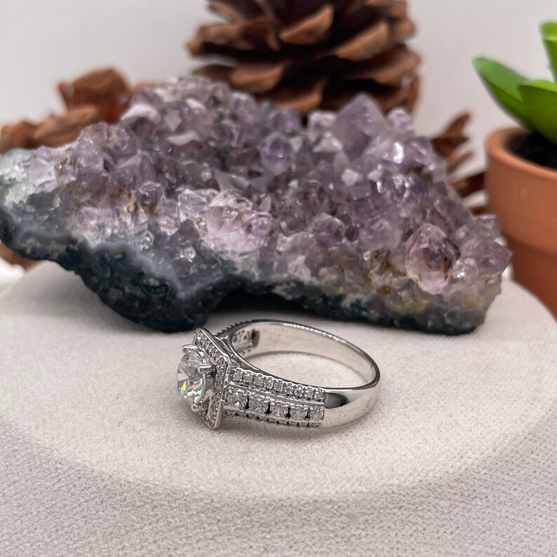May include: A silver ring with a large, clear, round gemstone surrounded by smaller gemstones. The ring has a double band with smaller gemstones set into the band.