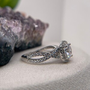 Puede incluir: Un anillo de plata con una piedra central grande, redonda y transparente rodeada de piedras m&aacute;s peque&ntilde;as. La banda tiene un dise&ntilde;o trenzado con piedras m&aacute;s peque&ntilde;as engastadas a lo largo de la banda.