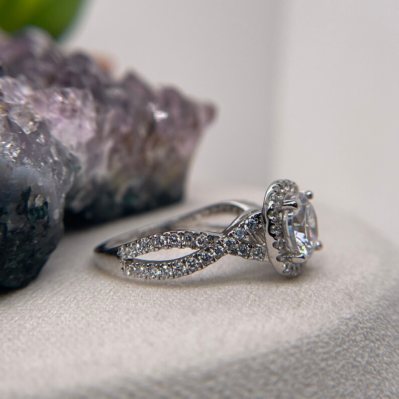 Puede incluir: Un anillo de plata con una piedra central grande, redonda y transparente rodeada de piedras m&aacute;s peque&ntilde;as. La banda tiene un dise&ntilde;o trenzado con piedras m&aacute;s peque&ntilde;as engastadas a lo largo de la banda.