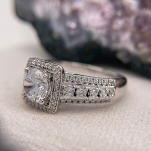 May include: A silver engagement ring with a large, round, clear center stone surrounded by smaller stones. The ring has a square-shaped band with a double row of smaller stones.