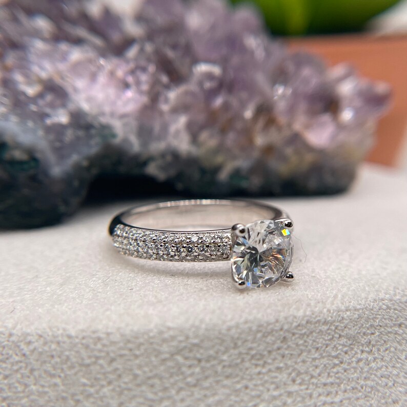 May include: A silver engagement ring featuring a large, round, clear diamond centre stone. The band is set with smaller diamonds in a pave setting.