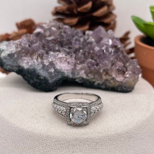 May include: A silver ring with a square-shaped center stone surrounded by smaller stones. The ring is set on a white surface with a purple amethyst crystal behind it.